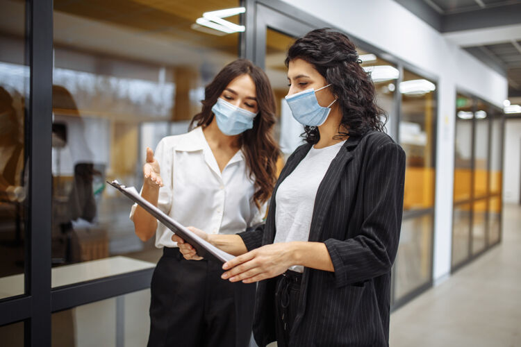 平板电脑两位女商人穿着医用无菌衣在办公室讨论工作问题面具工作在冠状病毒大流行隔离期间现代团队距离