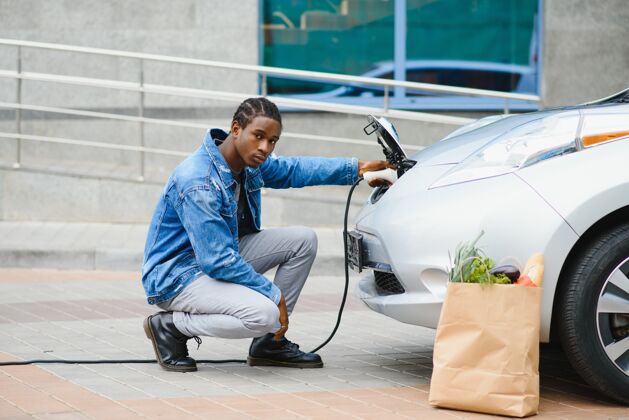 环境英俊的美国黑人男子坐在他的新的现代电动车旁边 拿着充电器的插头运输改变电力
