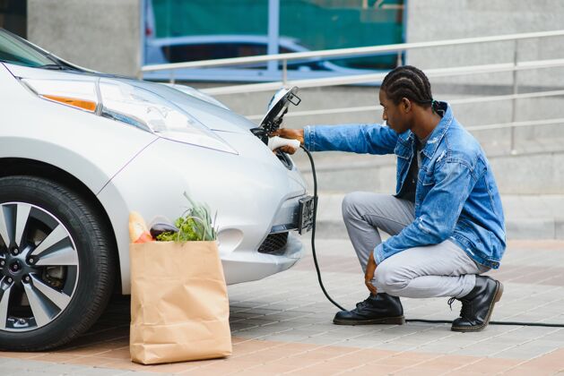 燃料英俊的美国黑人男子坐在他的新的现代电动车旁边 拿着充电器的插头电动车电源电池
