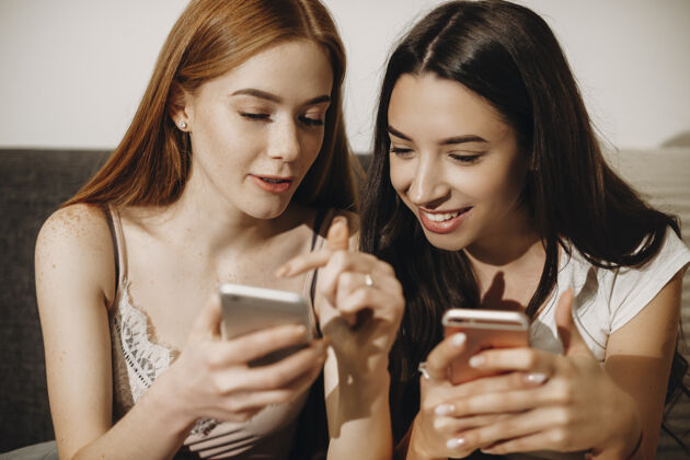 在线坐在沙发上 一位可爱的红发雀斑年轻女子在智能手机屏幕上向她的女性朋友展示的特写照片黑发女人小玩意