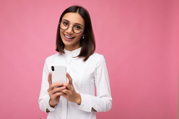 互联网迷人的微笑的年轻黑发女子身穿白色上衣和眼镜站在粉红色的墙上使用手机短信孤立电话移动手机