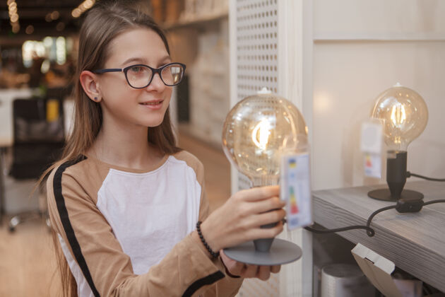 环境兴高采烈的少女手持灯泡 在家居用品店购物购买青少年女儿