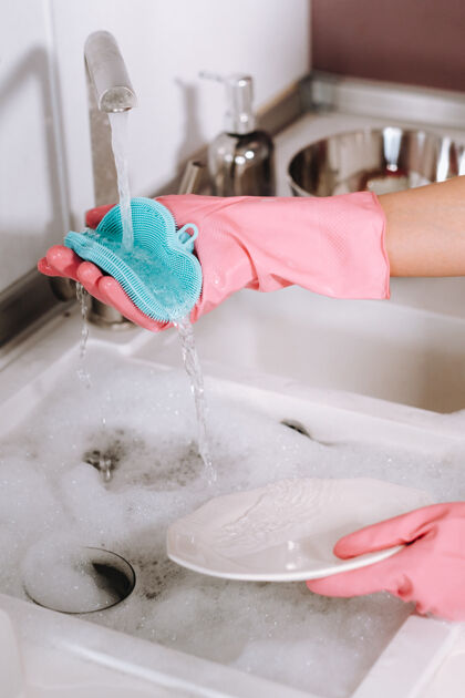 年轻人一个戴着粉色手套的家庭主妇在水槽里用手洗盘子清洁剂女孩在家里戴着手套洗碗盘子室内消毒