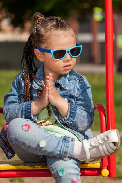 牛仔裤梳着辫子戴着太阳镜的时髦女婴坐在秋千上双手紧握牛仔黑发眼镜