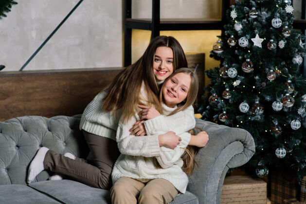 国际两个可爱的女孩在圣诞节摆出亲吻的表情树长-站在树旁的一位留着头发的欧洲女士和礼物孩子姐妹年轻