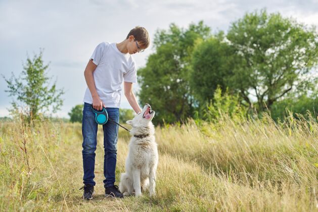 家庭户外白衣男孩画像谊少年带着宠物 少年漫步在夏日的草地上 风景如画的夕阳山水背景 复制空间友谊狗年轻
