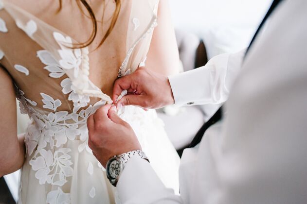 礼服一个穿着白衬衫 打着领带 戴着表的男人把扣子系在胸衣上礼服.新娘穿着蕾丝婚纱站在房间里优雅背部新郎