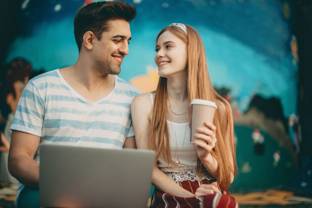 公园照片中 一对可爱的年轻夫妇面带微笑地看着对方 男人在笔记本电脑前工作 女人在公园里端着一杯咖啡约会浪漫朋友