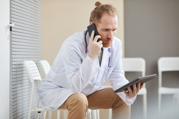 工作男医生坐在医院的椅子上用数字平板电脑和手机交谈互联网使用电话
