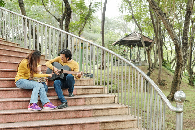 公园年轻女子指着丈夫手中的吉他讲解小儿子如何创作音乐女人台阶休闲服
