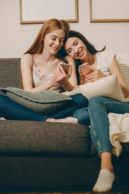 沙发两个美丽的年轻女子坐在家里的沙发上看着智能手机笑的照片社交舒适沙发
