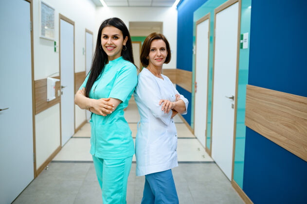 工人两位身着制服微笑的女医生在诊所摆姿势走廊.专业医院的医学专家 喉科医生或耳鼻喉科医生 妇科医生或乳腺科医生 外科医生医院室内工作