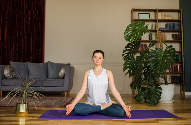运动在家里的一个房间里 一个穿着运动制服的瑜伽女士坐在垫子上的莲花姿势姿势地板房子