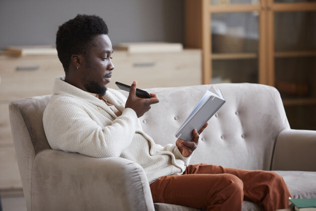 家庭生活坐在沙发上看书 用手机录下语音信息的非洲男人他坐在房间里电脑家庭房间舒适