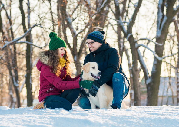 猎犬在阳光明媚的日子里 一对可爱的年轻夫妇和他们的金毛猎犬在冬季公园里玩得很开心微笑女人在冬天的雪乡 男人抱着狗男性冬天玩耍