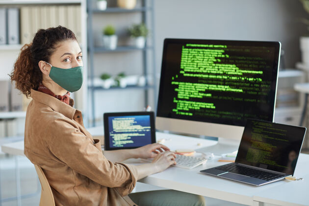 防护戴着防护面具的年轻程序员坐在办公桌前看着前方 桌上放着电脑计算机黑客流行病持有
