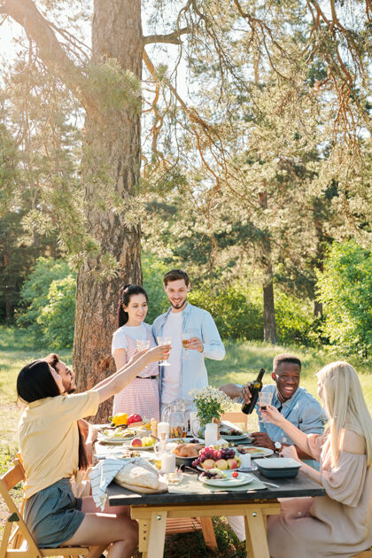 夏天三对年轻的多元文化情侣在丰盛的节日餐桌上碰杯 一边在松树下享用户外晚餐男孩孩子烧烤