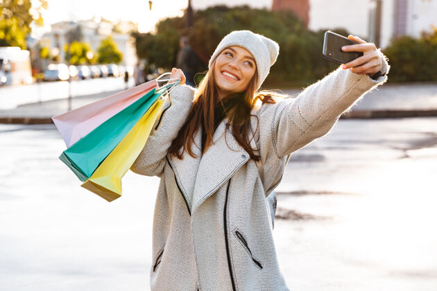 年轻一个红发快乐的女人拿着购物袋走在户外用手机自拍的照片自拍微笑举行