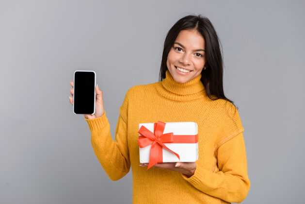 漂亮一个快乐随意的年轻女子的肖像被隔离在灰色的墙上 显示手机屏幕空白 手持礼品盒购物空白休闲