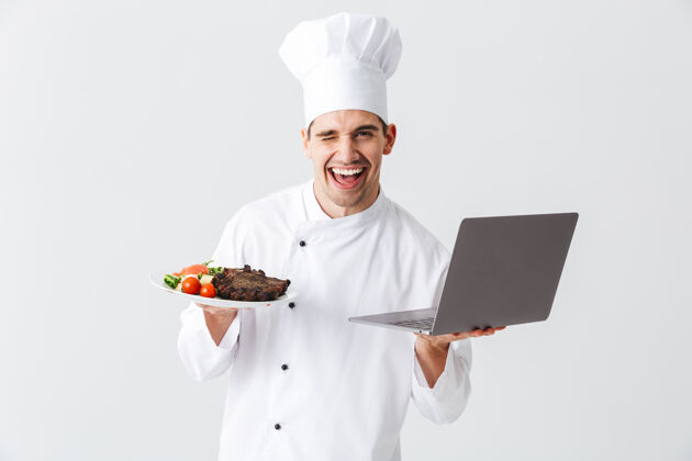工作兴奋的厨师穿着制服站在白色的墙上 拿着笔记本电脑 展示着一道菜笔记本电脑职业男性