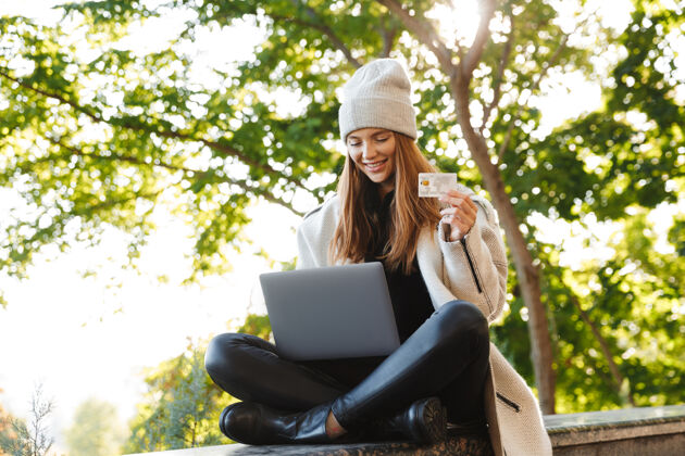 成功快乐的年轻女子穿着秋大衣 戴着秋帽坐在户外 用笔记本电脑 展示塑料信用卡外套秋天购买