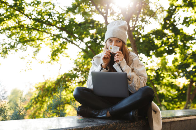 使用快乐的年轻女子穿着秋大衣 戴着秋帽坐在户外 用笔记本电脑 展示塑料信用卡购物付款微笑