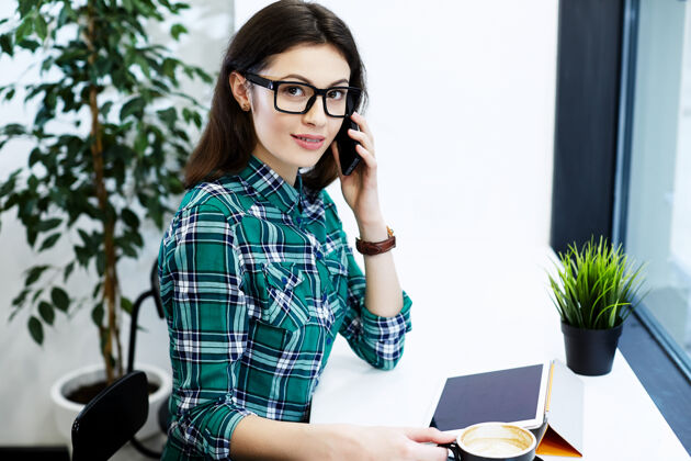 工作黑头发的漂亮女孩穿着衬衫戴着眼镜坐在咖啡馆里拿着平板电脑和一杯咖啡 讲着手机 自由职业者的概念 看着相机青少年杯子女性
