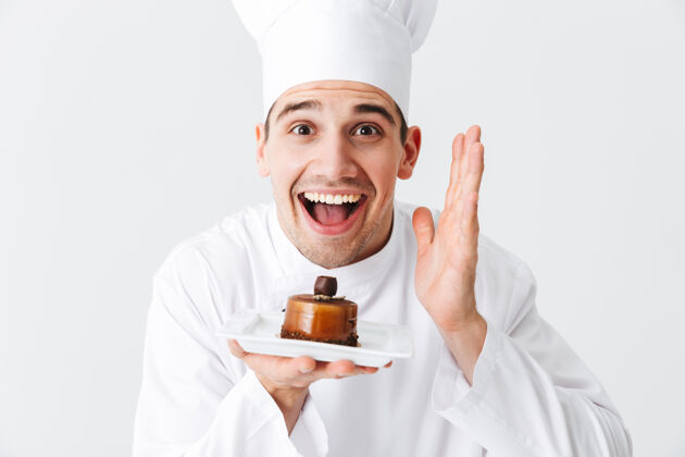 制造开朗的厨师穿着制服 在白色的墙上展示一个盘子上的糕点帽子制服面包房
