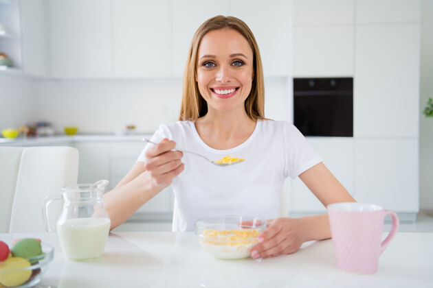 女人照片中美丽的主妇端着满满一勺鲜奶早餐玉米片美味健康的吃着享受美味的饭菜坐在餐桌上白光厨房里家庭主妇维生素吃