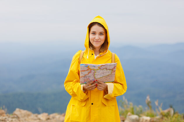 游客穿着黄色雨衣的时髦女孩背着背包在山顶上看地图旅行年轻观光