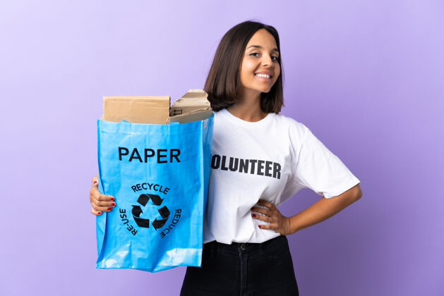 积极一个年轻的拉丁女子拿着一个装满废纸的回收袋 在紫色的孤零零地摆姿势 双臂放在臀部 面带微笑女性循环环境