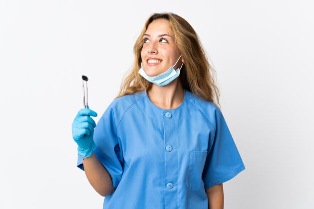 成年人女牙医拿着工具孤立地站在白纸上一边想一边抬头看健康人女人