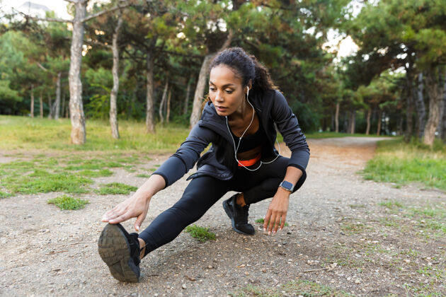 锻炼一位20多岁的美籍黑人妇女穿着黑色运动服 在绿色公园里锻炼身体 伸展双腿女性音乐女运动员