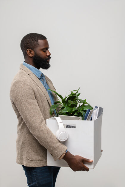 包一位留着胡须的年轻的非裔美国上班族的侧视图 他正装在一个装有耳机 绿色植物和其他用品的手提箱里商业人年轻人