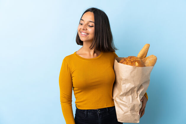 碳水化合物一个年轻的拉丁女人买了一些孤立在蓝色的面包 面带微笑地看着旁边面包店面包房黑发