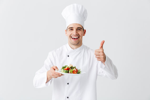 菜肴兴奋的男厨师穿着制服 在白色墙壁上展示一盘新鲜的蔬菜沙拉 竖起大拇指欢呼烹饪工作
