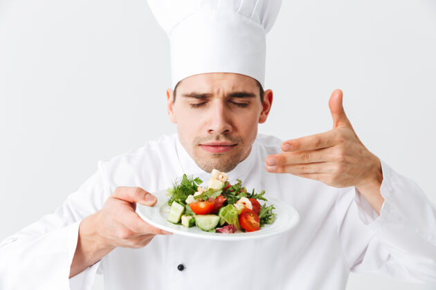准备兴奋的男厨师穿着制服 把新鲜的蔬菜沙拉放在隔离在白色墙上的盘子上举行欢呼成人