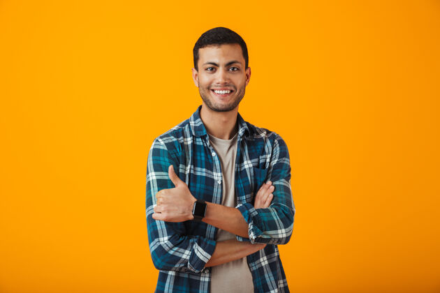 人一个穿着格子衬衫的快乐的年轻人孤零零地站在橙色的墙上 双臂交叉着格子年轻健身