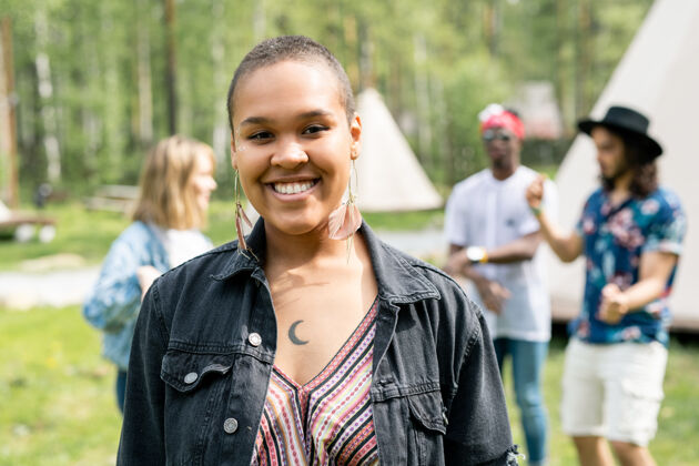 欢乐一个戴着皮耳环微笑的非洲裔美国女孩站在营地的朋友面前的画像团聚一群人牙齿微笑