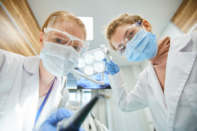 医院两个戴着防护面具和眼镜的牙医在团队中使用医疗器械时注视前方的肖像室内实验室外套诊所