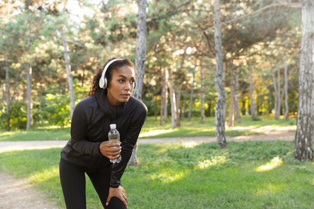 温暖20多岁的女性穿着黑色运动服 戴着耳机 拿着水瓶在绿色公园散步运动员年轻自然