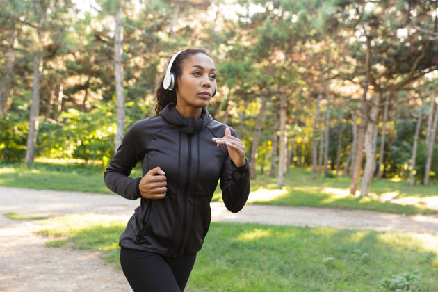 运动一个20多岁的精力充沛的女人穿着黑色运动服 戴着耳机 在绿色公园里跑步锻炼深色头发早晨
