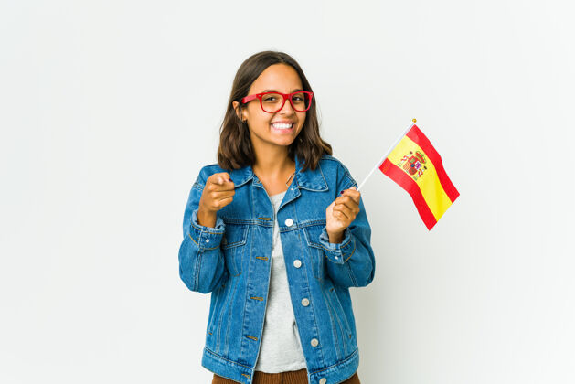 观点年轻的西班牙女子举着一面隔离在白墙上的国旗 微笑着指着前方高兴人高兴