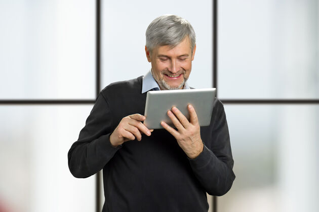 电脑成熟的男人微笑着看平板电脑上的新闻快乐的老人看着pc平板电脑 微笑着看窗户商务男性工人