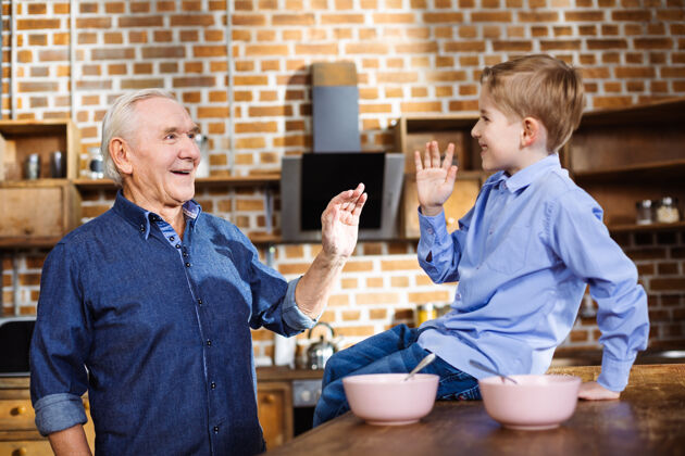 孙子快乐的老人站在厨房里和他的孙子玩大笑桌子谷类食品
