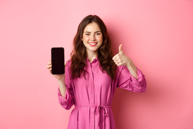 女性科技概念穿着裙子的年轻女孩 展示她的智能手机屏幕 竖起大拇指 点头表示赞同 称赞好的应用程序 站在粉色的墙上时尚电话兴奋