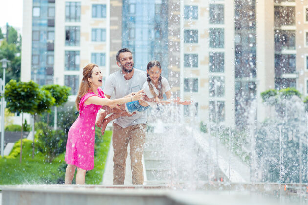 关怀快乐的时光快乐的父母站在喷泉边和他们的女儿玩耍关系积极休息日