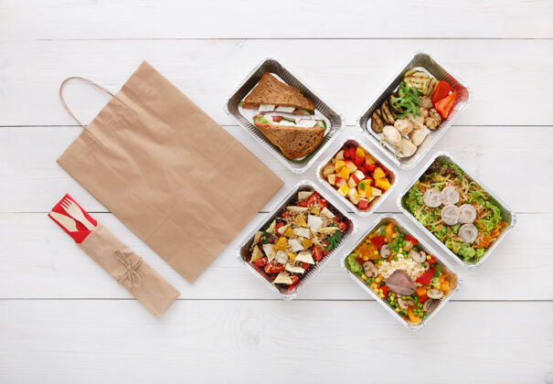餐厅健康食品配送外卖食品铝箔盒中的蔬菜 肉类和水果 餐具 水和牛皮纸包装俯视图 平放在白色木头上 有复印空间美味自然平铺