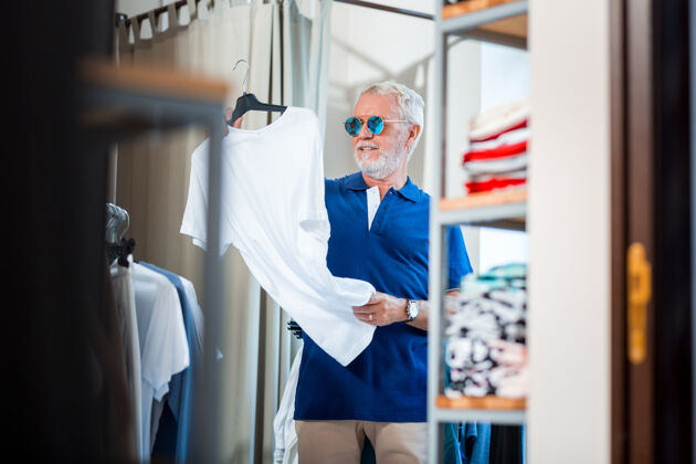 时尚新衣服好奇的老顾客腰部向上看白色t恤 同时用双手握住它购买老年人休息