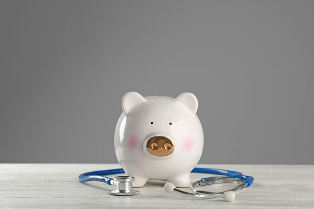 银行可爱的小猪存钱罐和听诊器放在灰色背景下的木桌上昂贵存款金融